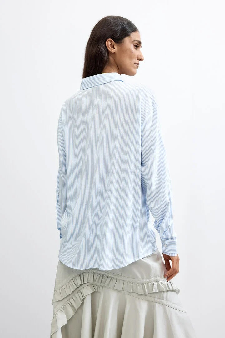 A woman shown from the back wearing a light stick blue and white vertically striped button-up shirt and a light beige ruffled skirt, set against a plain white studio background.