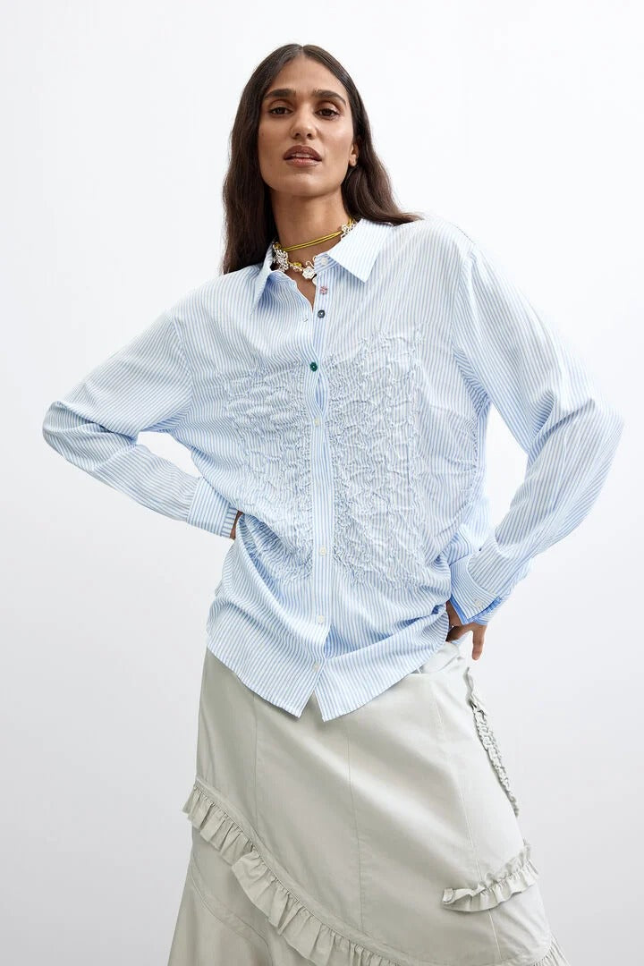 A woman modeling a light stick blue and white vertically striped long-sleeve button-up shirt with intricate embroidered detailing on the front, paired with a light beige skirt, against a plain white studio background.