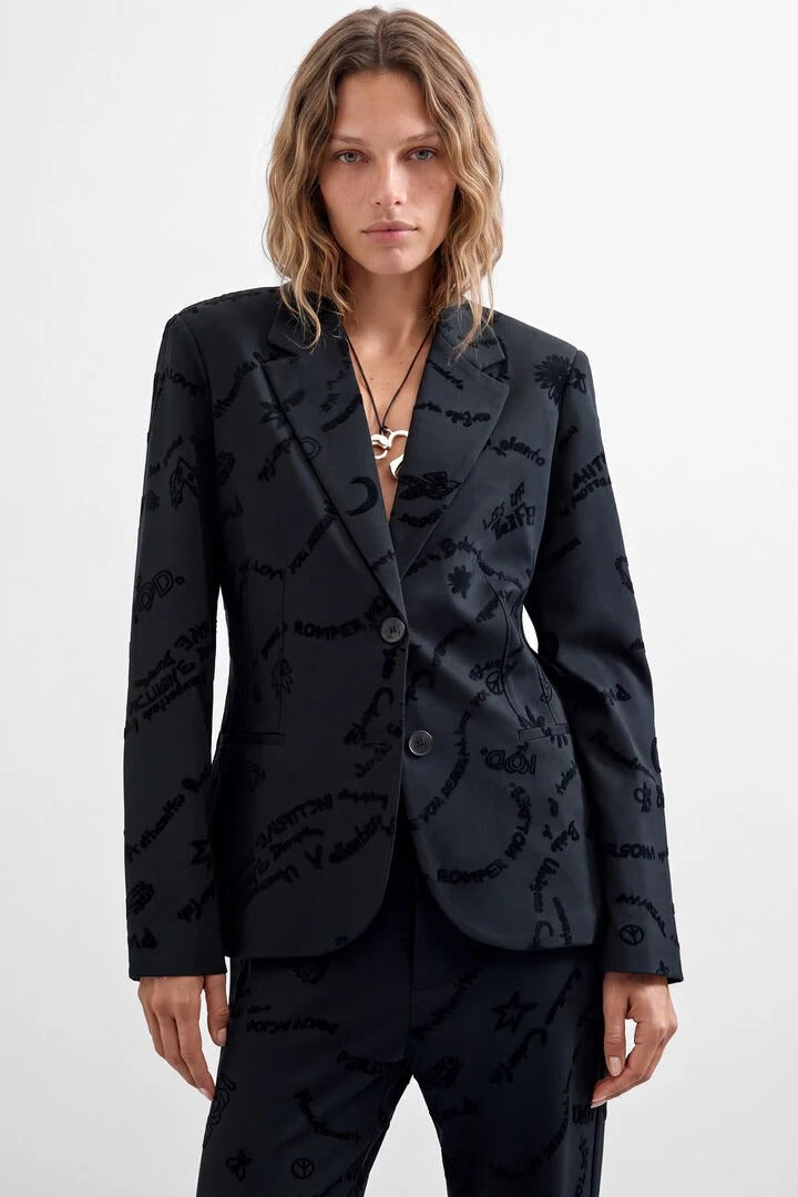 A woman modeling a black tailored blazer and matching trousers with an allover tonal script pattern, photographed against a clean light gray studio background.