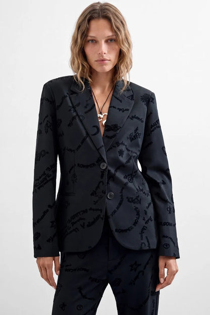 A woman modeling a black tailored blazer and matching trousers with an allover tonal script pattern, photographed against a clean light gray studio background.