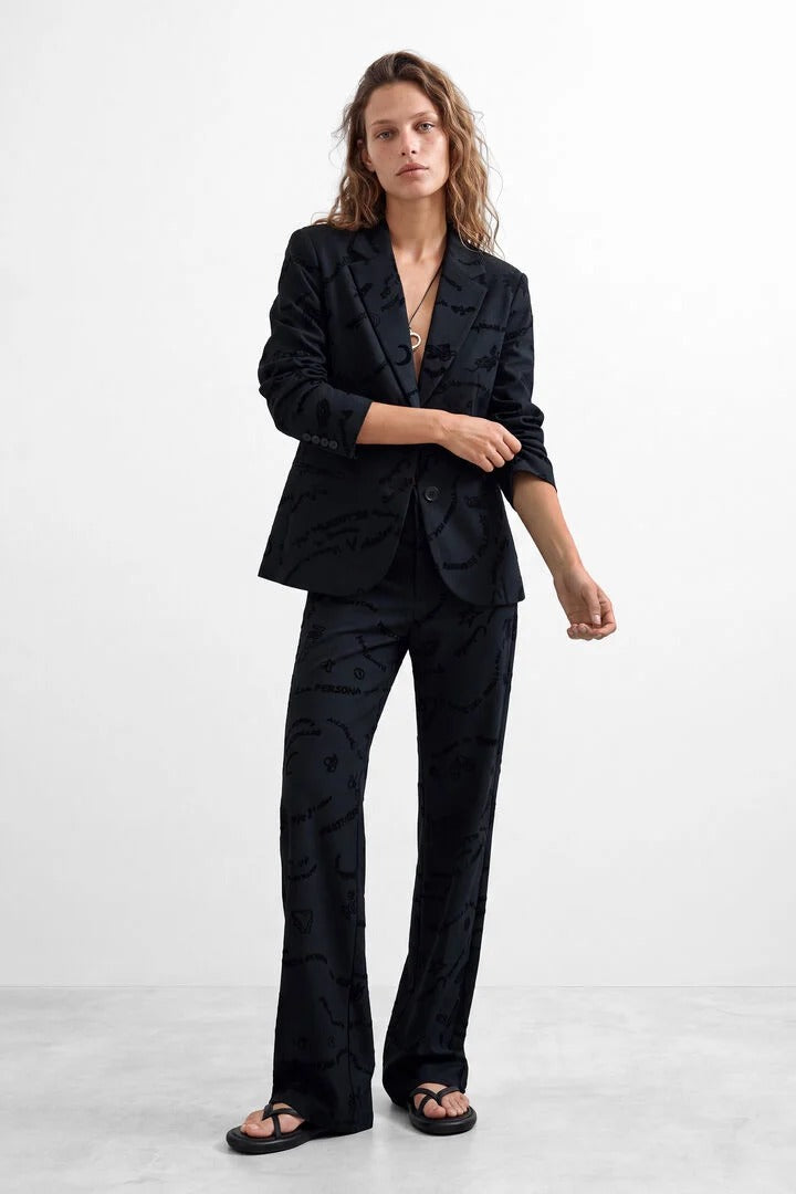 A woman modeling a black two-piece suit with a subtle tonal abstract pattern, featuring a cropped blazer and matching wide-leg trousers, styled with black sandals against a light gray studio background.