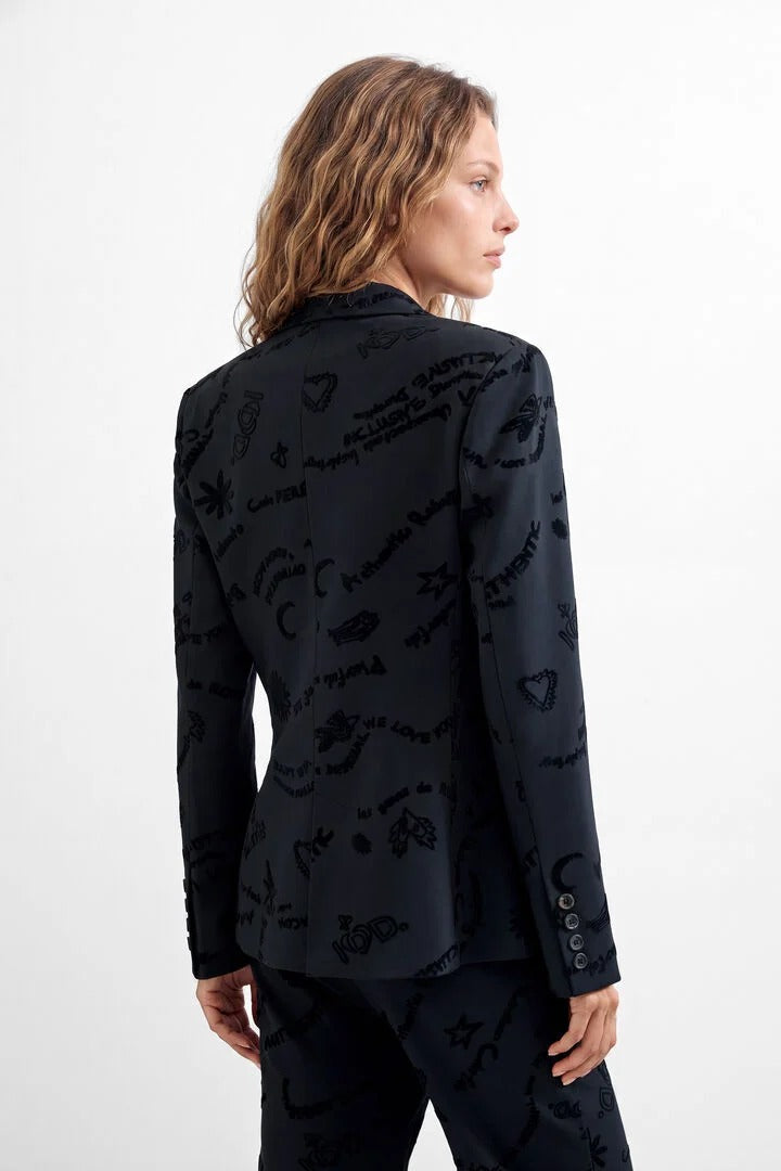 A woman shown from the back wearing a black blazer and matching trousers with an allover tonal graphic print of handwritten text and symbols, against a plain white studio background.