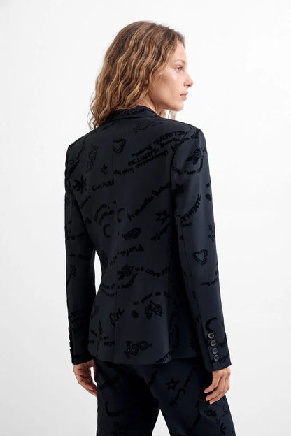 A woman shown from the back wearing a black blazer and matching trousers with an allover tonal graphic print of handwritten text and symbols, against a plain white studio background.