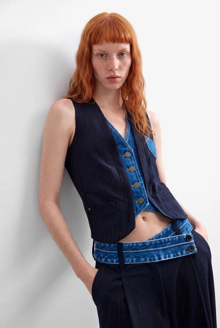 A model with red hair wearing a dark navy azul oscuro double lapel pinstripe vest over a blue denim button-front top, paired with matching dark navy pinstripe trousers and a blue denim belt, posing against a light gray wall.