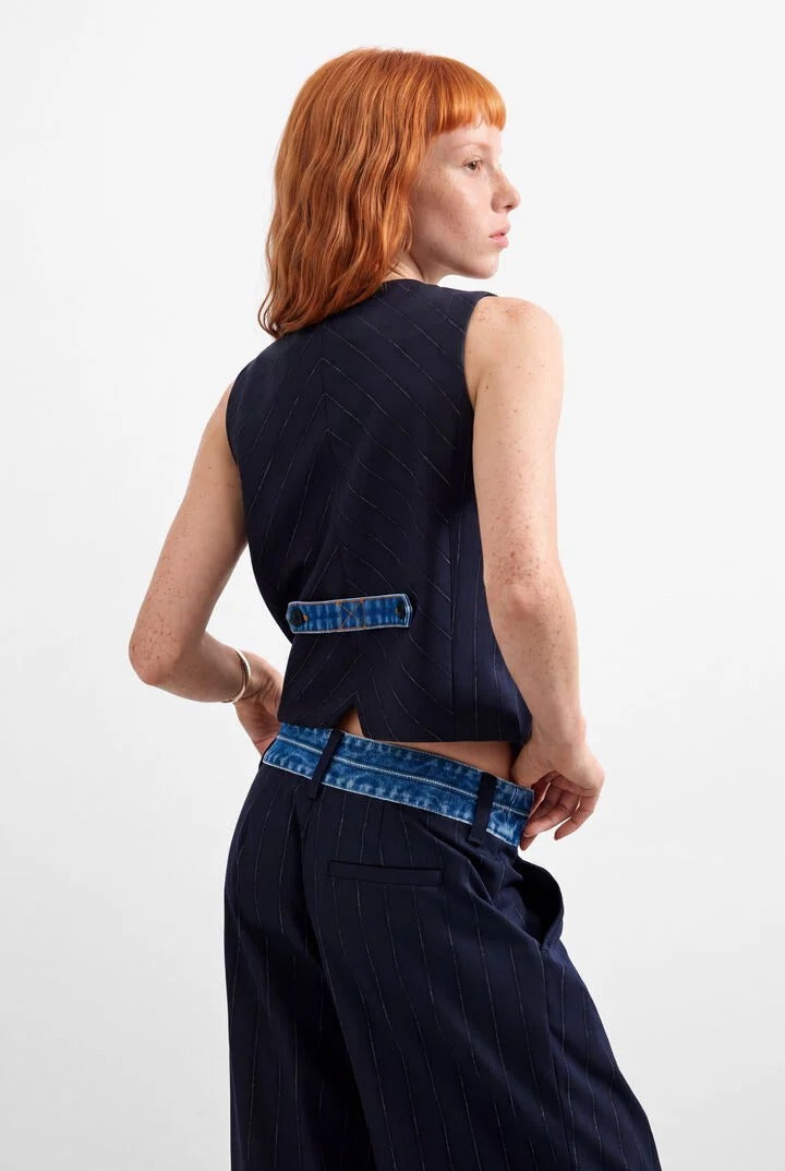 A model with red hair is shown from the back wearing a azul oscuro dark navy sleeveless vest with a blue denim patch detail and matching navy pinstripe trousers with a denim belt, against a plain white studio background.