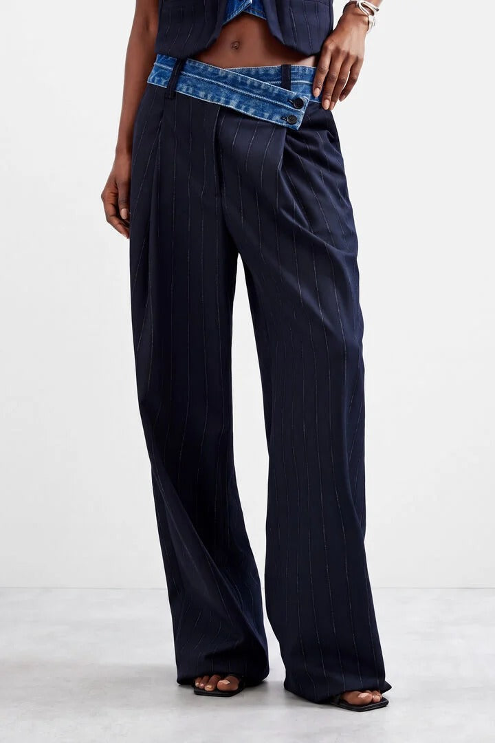 A model wearing dark navy wide-leg pinstripe trousers with a contrasting blue denim waistband, paired with a dark top and black sandals, against a light gray studio background.