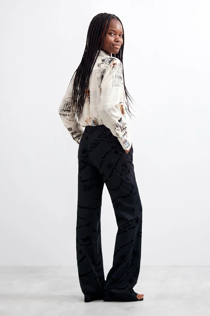 A person wearing black wide-leg long printed trousers with a tonal black-on-black script and heart pattern, paired with a light patterned top and brown sandals, shown against a plain white studio background.