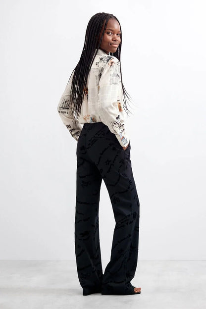 A person wearing black wide-leg long printed trousers with a tonal black-on-black script and heart pattern, paired with a light patterned top and brown sandals, shown against a plain white studio background.