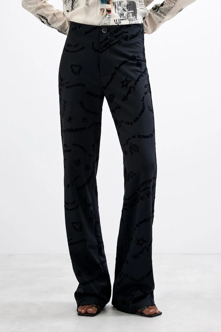 A person wearing black wide-leg trousers with a tonal black-on-black script and heart pattern, paired with a light patterned top and brown sandals, shown against a plain white studio background.