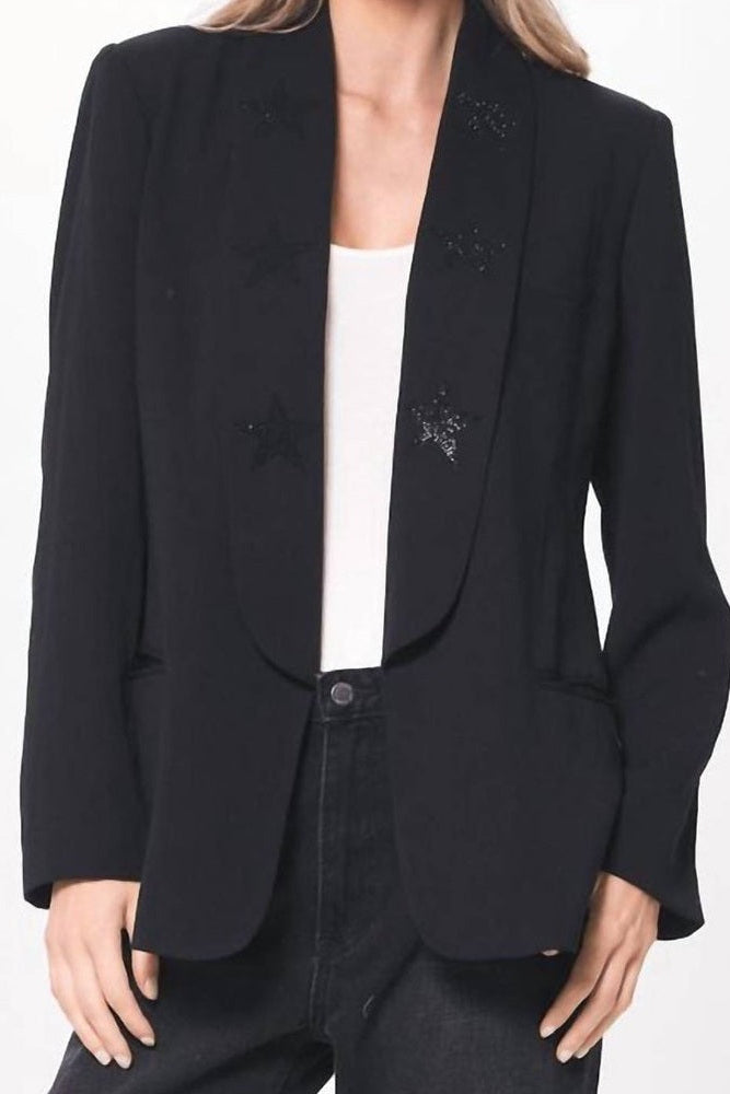 A woman wearing a black blazer with star-shaped sequin embellishments on the lapels, styled over a white top and dark jeans against a plain white background.