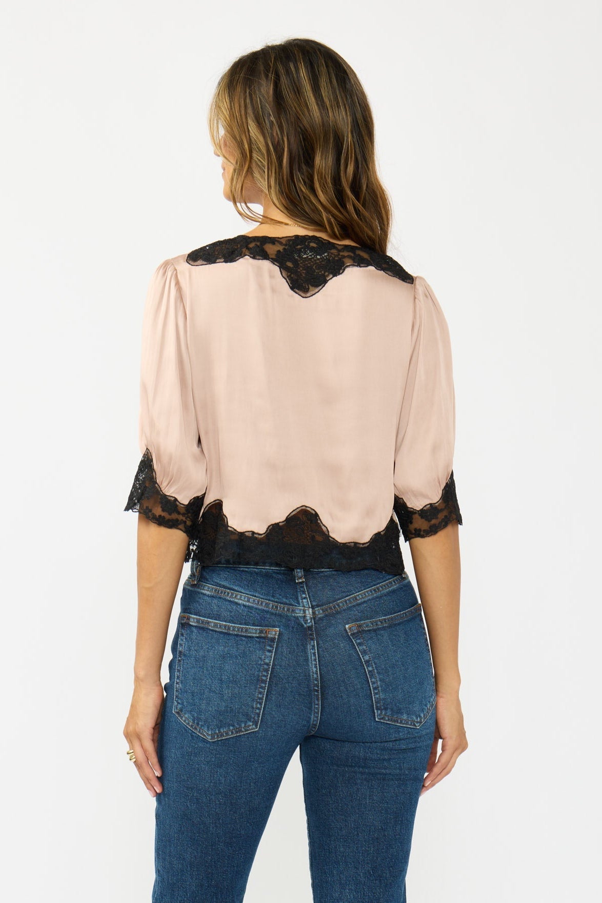 A woman viewed from the back wearing a champagne blouse with black lace trim at the shoulders, neckline, and hem, paired with blue jeans against a plain white studio background.