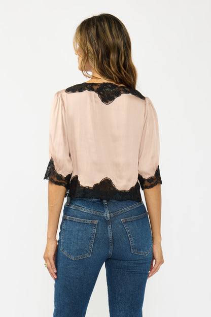 A woman viewed from the back wearing a champagne blouse with black lace trim at the shoulders, neckline, and hem, paired with blue jeans against a plain white studio background.