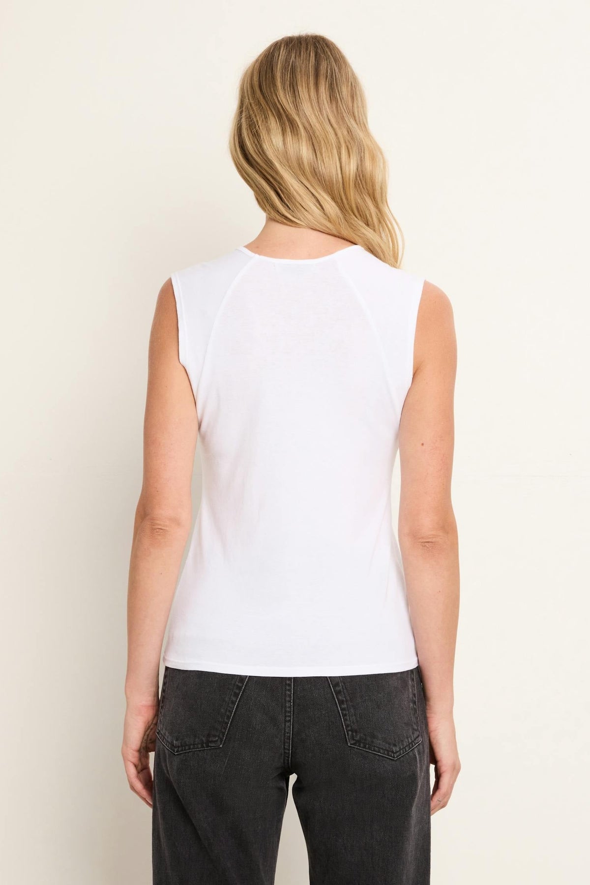A woman viewed from the back wearing a white sleeveless top and dark gray jeans against a plain off-white studio background.