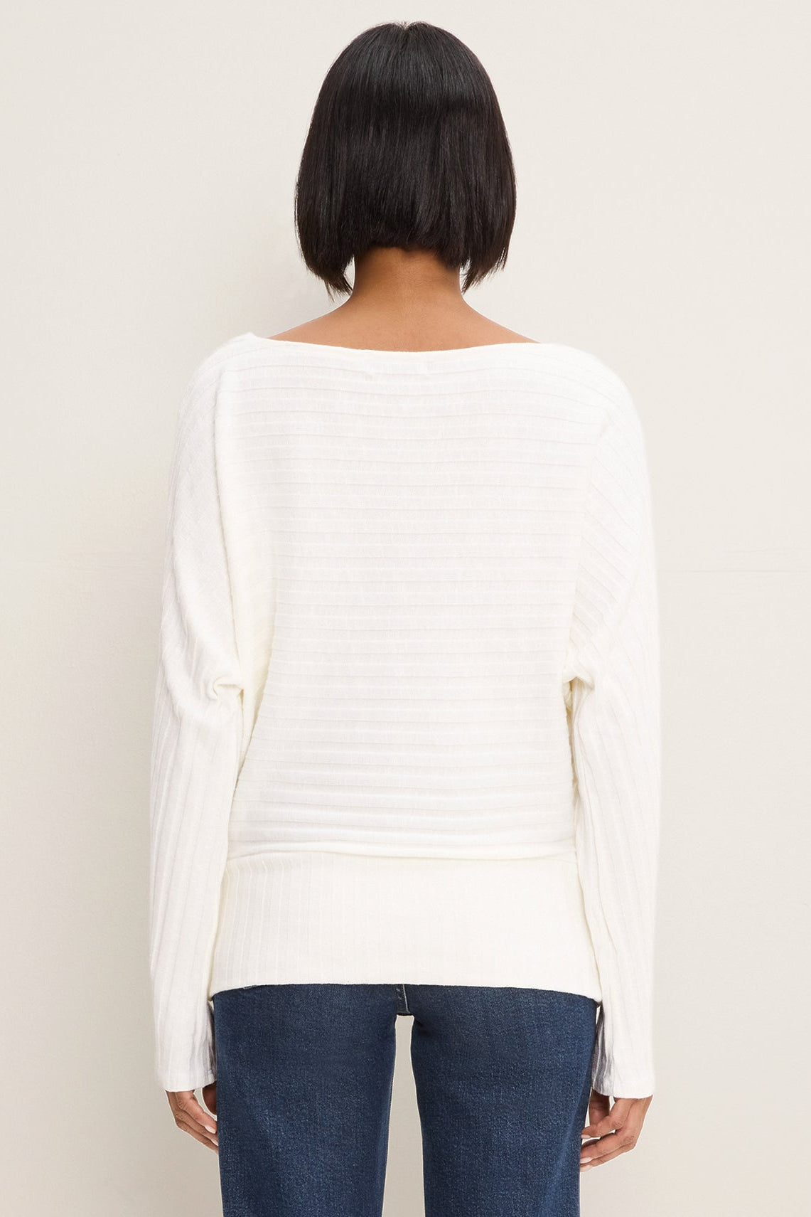 A woman viewed from the back wearing a coconut milk colored ribbed knit sweater with a wide boat neckline and long sleeves, paired with dark blue jeans, against a plain light beige background.