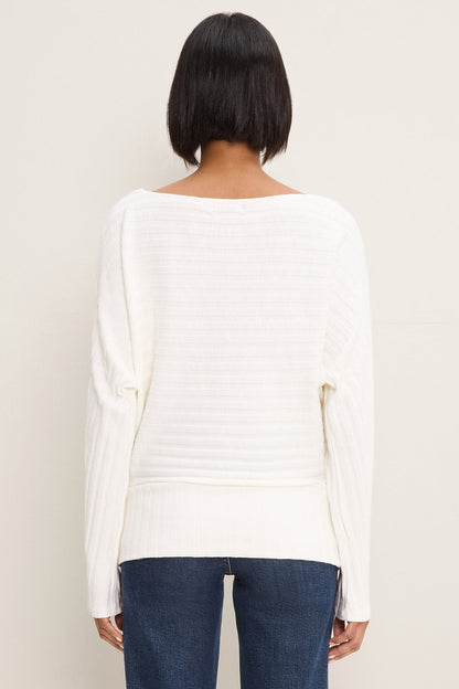 A woman viewed from the back wearing a coconut milk colored ribbed knit sweater with a wide boat neckline and long sleeves, paired with dark blue jeans, against a plain light beige background.