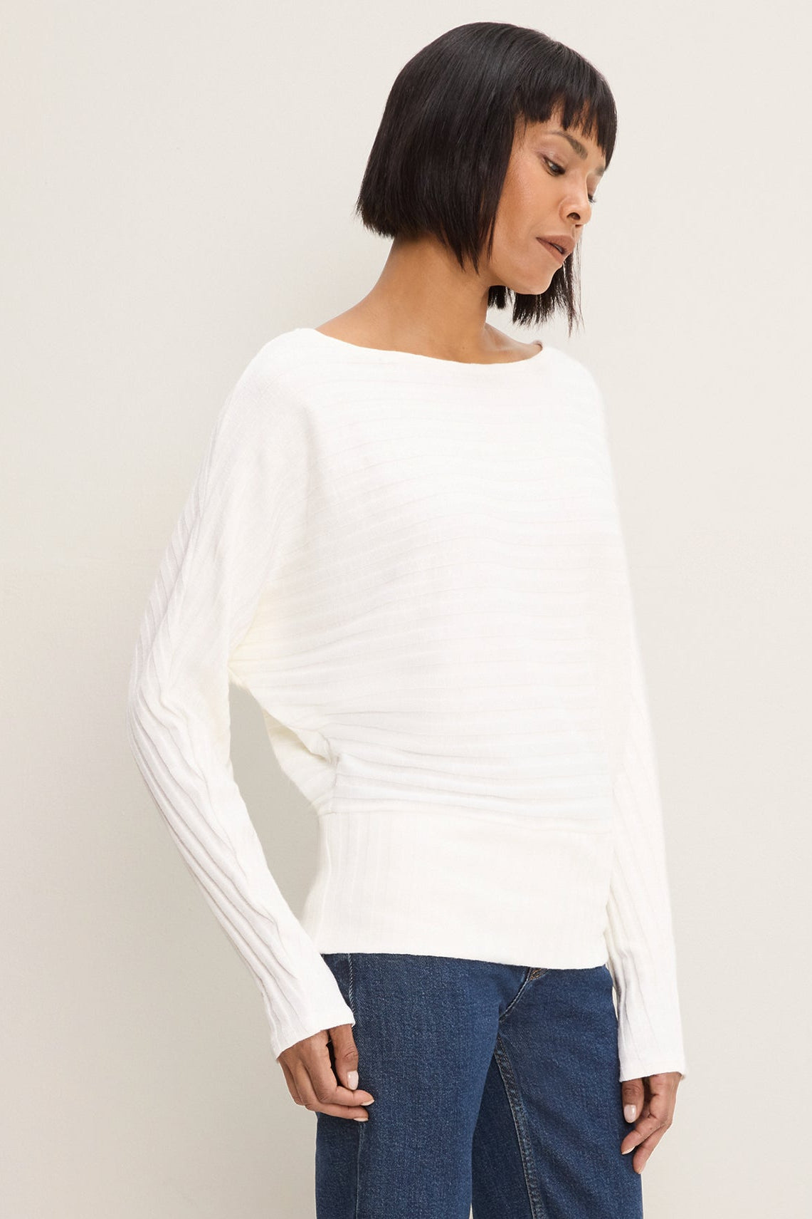 A woman wearing a coconut milk colored ribbed knit long-sleeve boat-neck sweater and blue jeans, photographed against a plain light beige studio background.