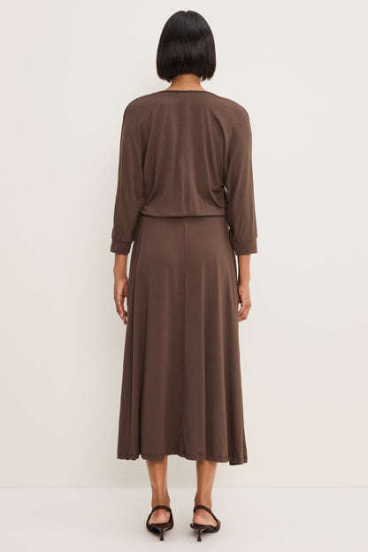 A woman from the back  modeling a Molasses colored long-sleeve V-neck midi dress, paired with brown pointed-toe heels against a plain off-white studio background.