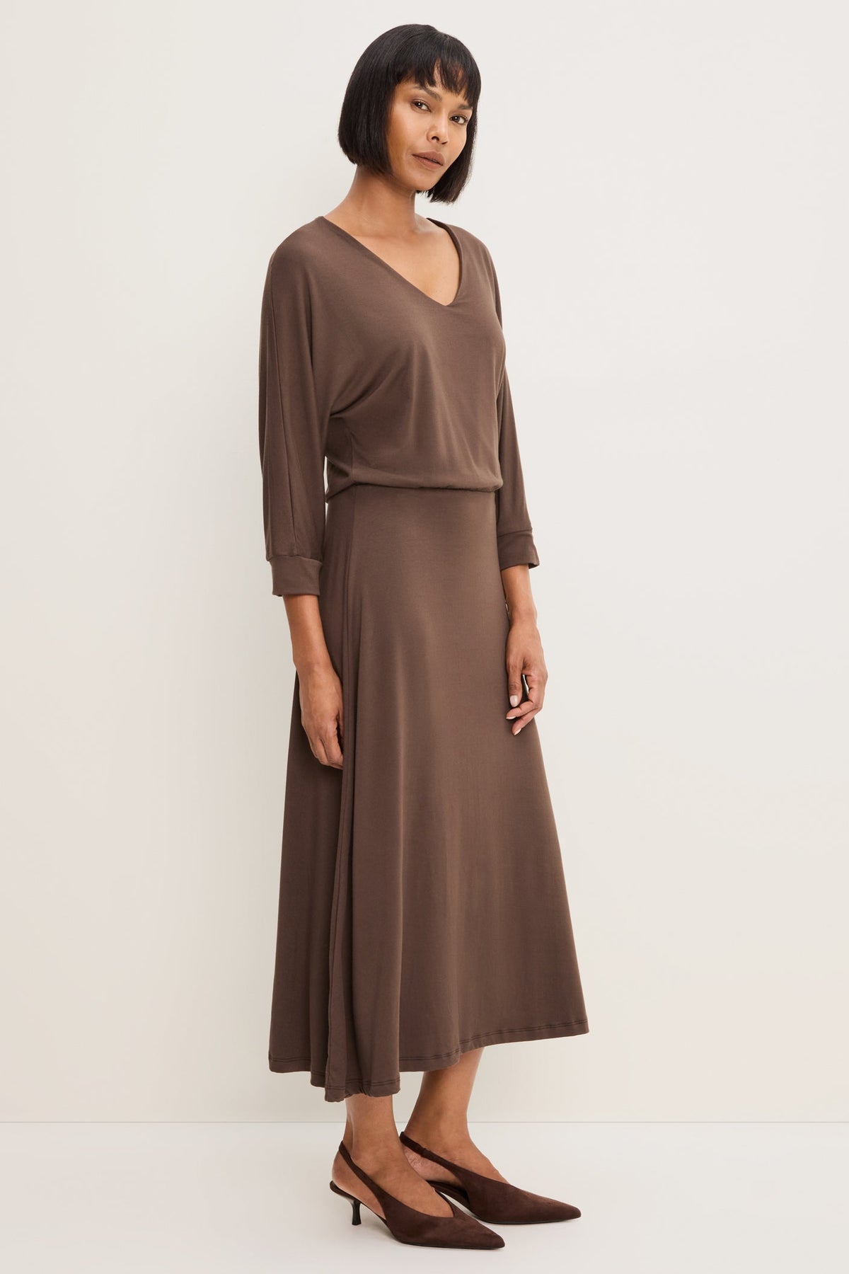 A woman modeling a Molasses colored long-sleeve V-neck midi dress, paired with brown pointed-toe heels against a plain off-white studio background.