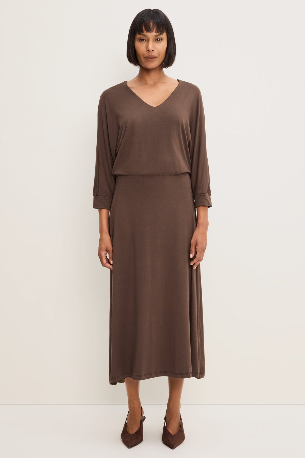 A woman modeling a Molasses colored long-sleeve V-neck midi dress, paired with brown pointed-toe heels against a plain off-white studio background.