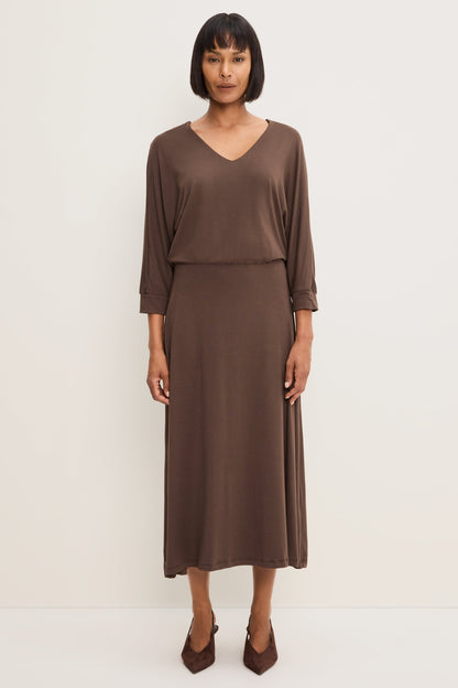 A woman modeling a Molasses colored long-sleeve V-neck midi dress, paired with brown pointed-toe heels against a plain off-white studio background.