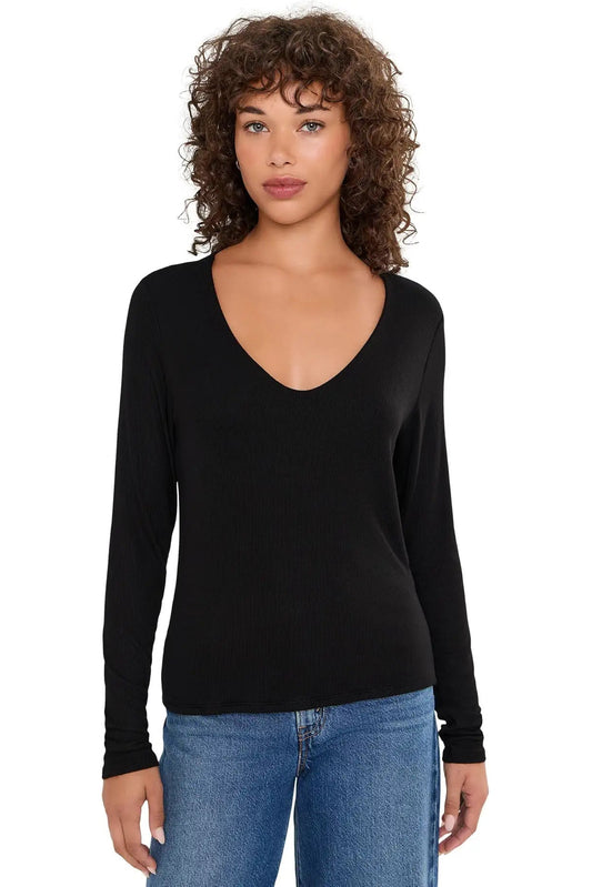 A woman with curly brown hair wearing a black long-sleeve V-neck ribbed knit top and blue jeans, standing against a plain white background.