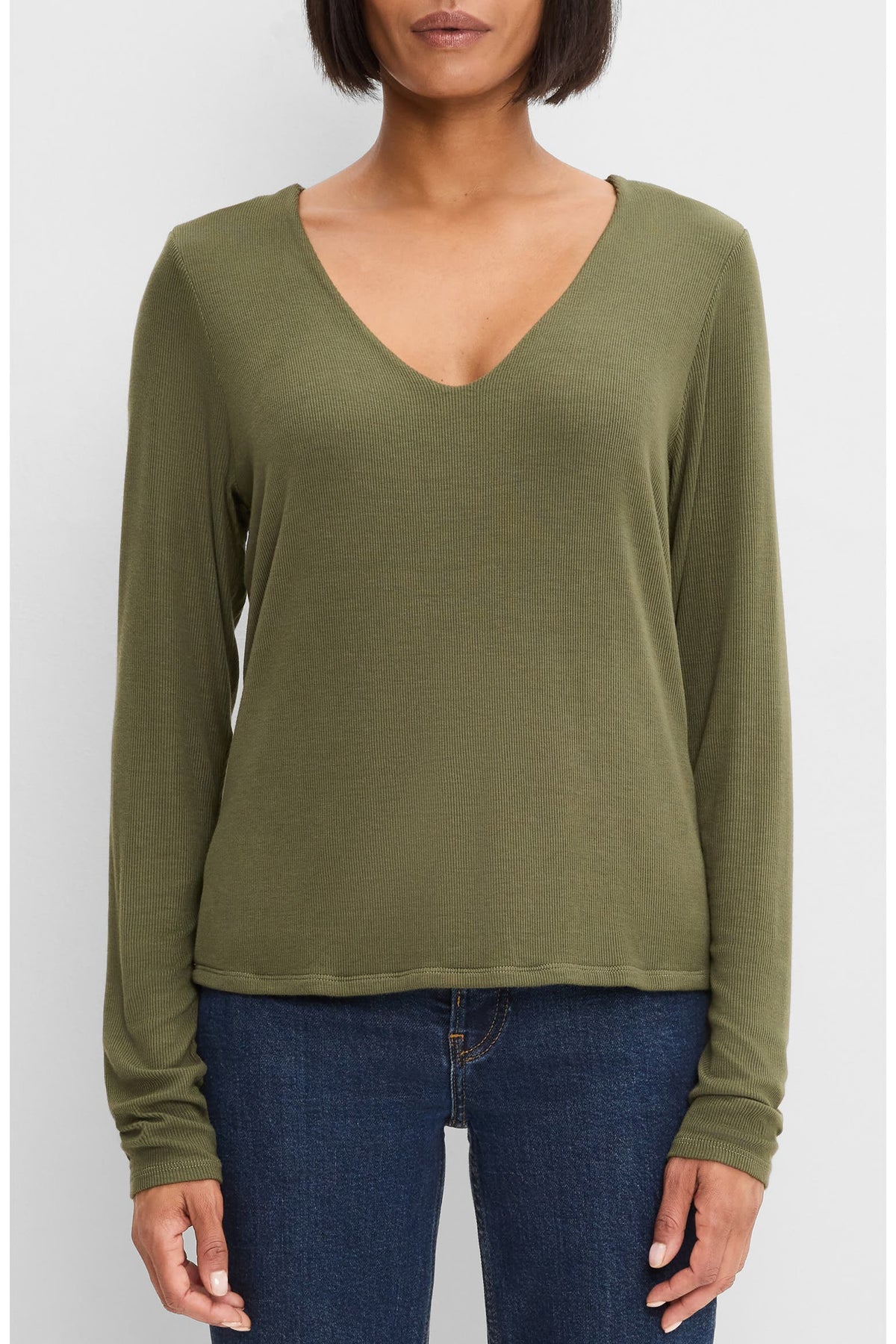A woman wearing a long-sleeve dark basil green V-neck top and dark blue jeans, shown from the chin down against a light gray studio background.