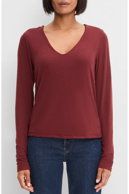 A woman wearing a long-sleeve V-neck top in a port royale color, paired with dark blue jeans, against a light gray studio background.