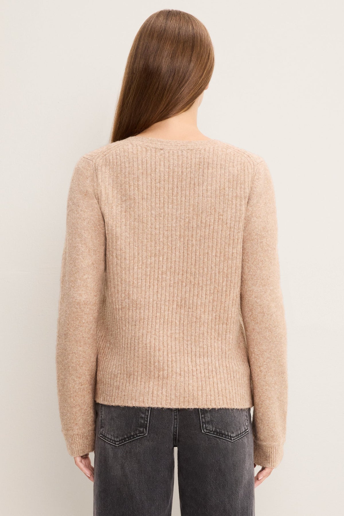 A woman viewed from the back wearing an oatmeal ribbed knit sweater and dark gray jeans, set against a plain off-white background.