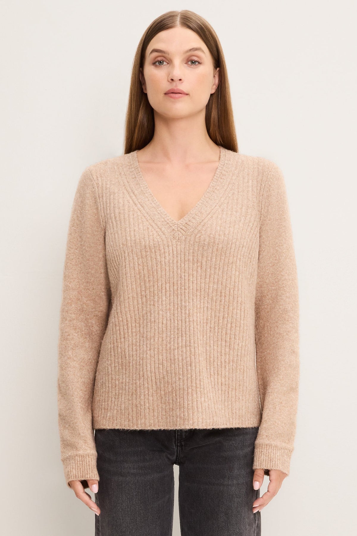 A woman modeling an oatmeal colored ribbed knit V-neck sweater with long sleeves, paired with dark gray jeans, against a plain off-white studio background.
