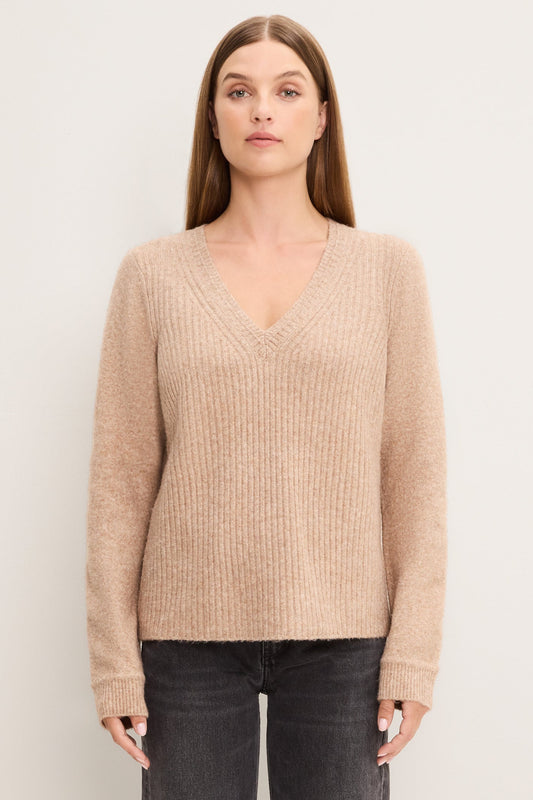A woman modeling an oatmeal colored ribbed knit V-neck sweater with long sleeves, paired with dark gray jeans, against a plain off-white studio background.
