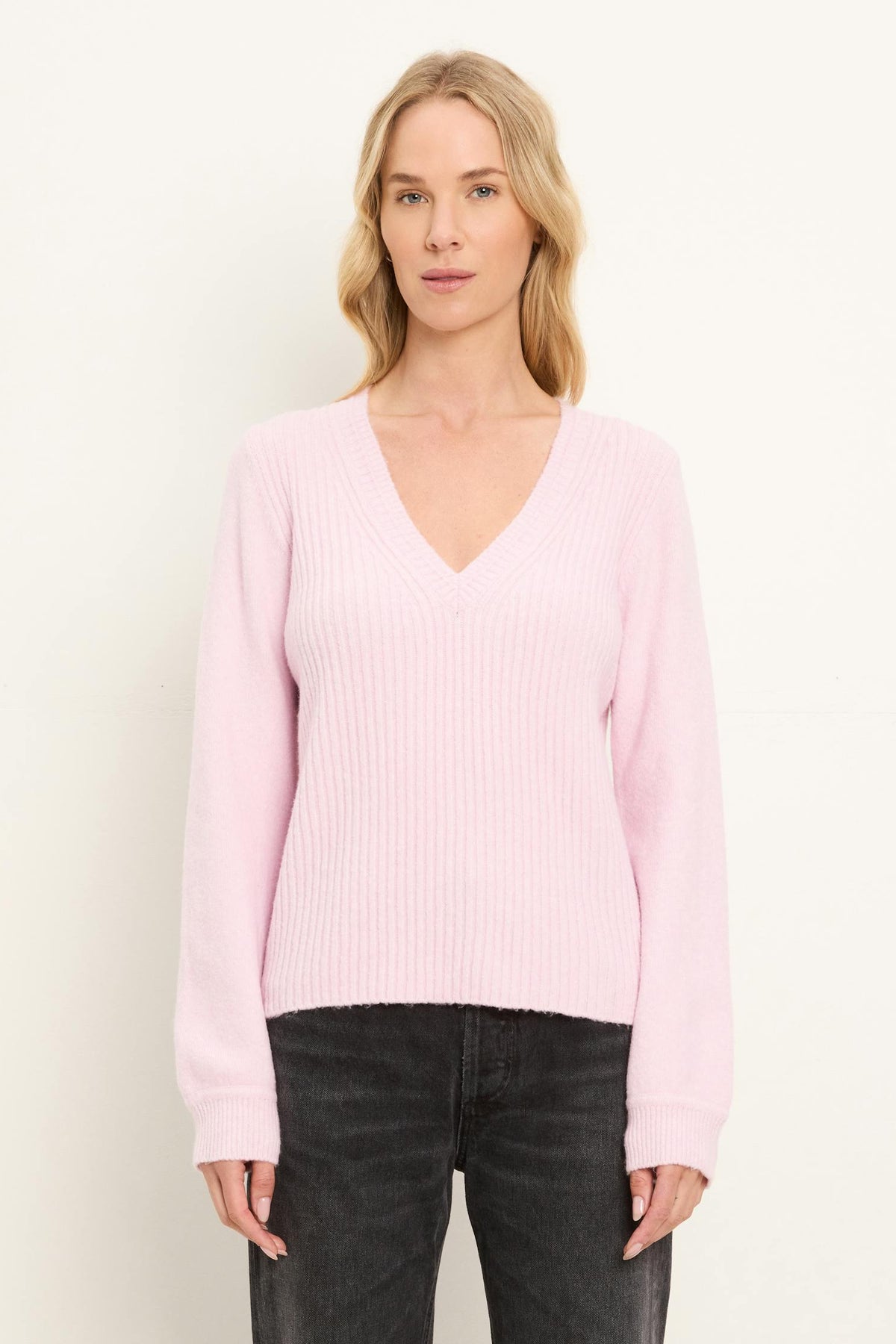 A woman shown from the front wearing a pink tulip colored v neck sweater and washed black jeans on  a white background.