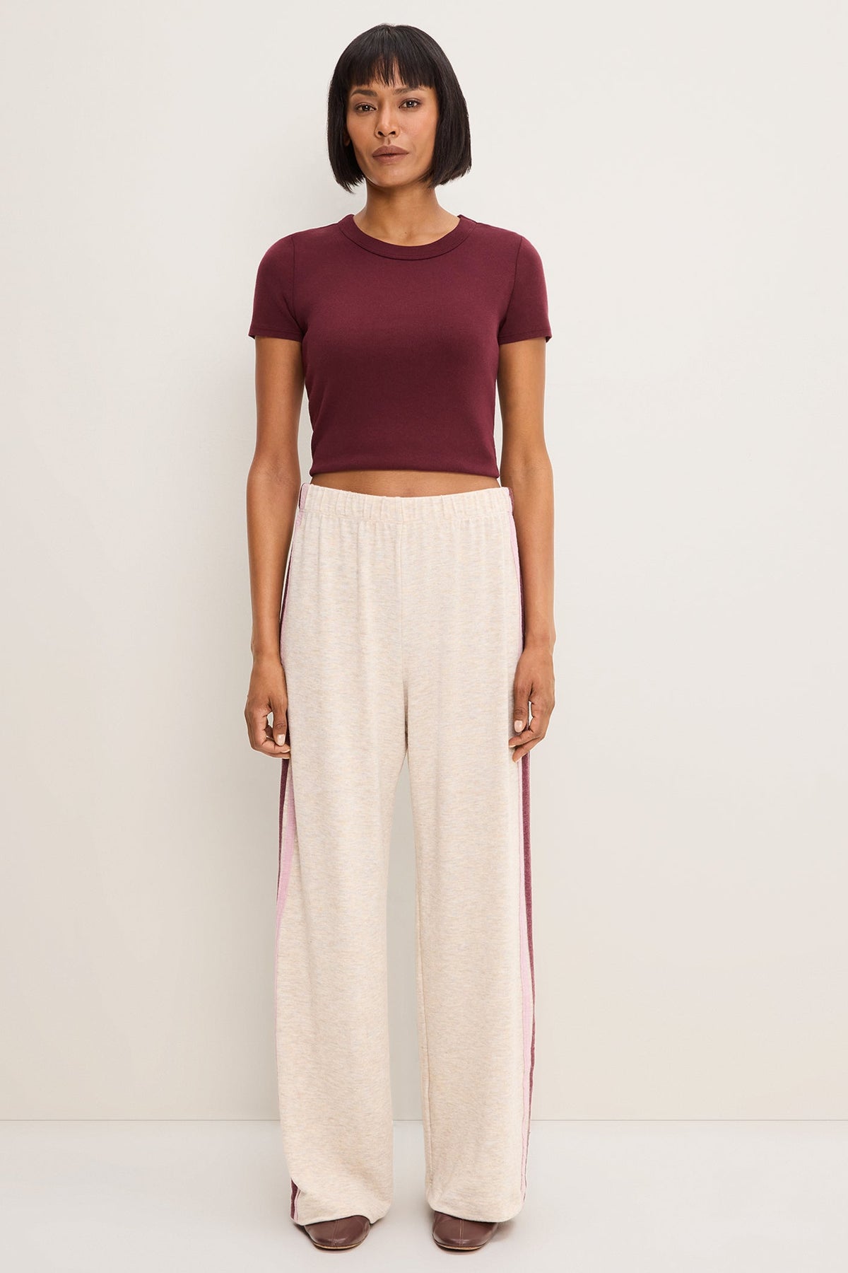 A model stands against a plain off-white studio background wearing a short-sleeve crewneck crop top in deep burgundy and wide-leg cropped pants in heather oatmeal with burgundy side stripes.