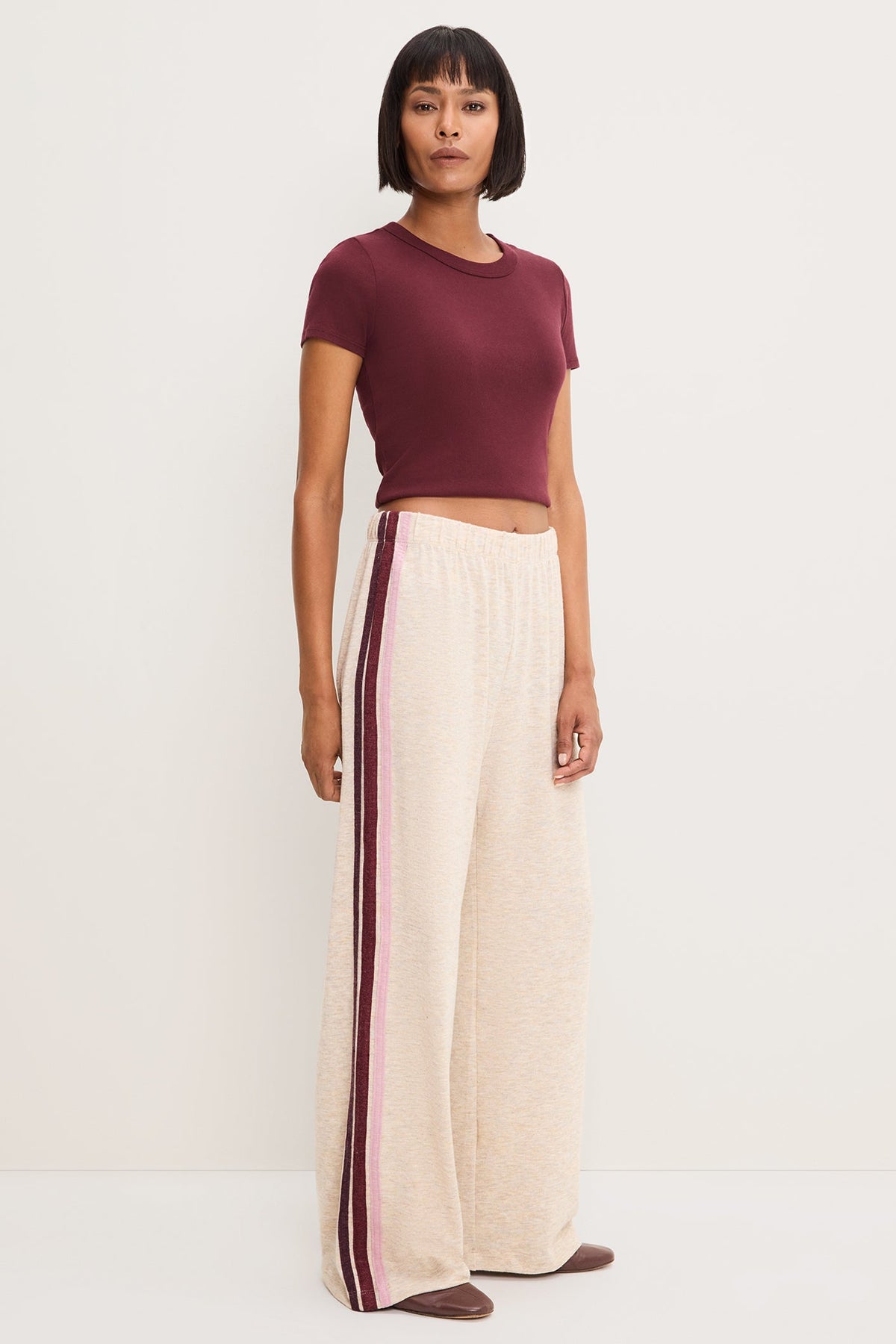 A model stands against a plain off-white studio background wearing a short-sleeve maroon crop top and wide-leg heather oatmeal pants with a burgundy and pink side stripe.