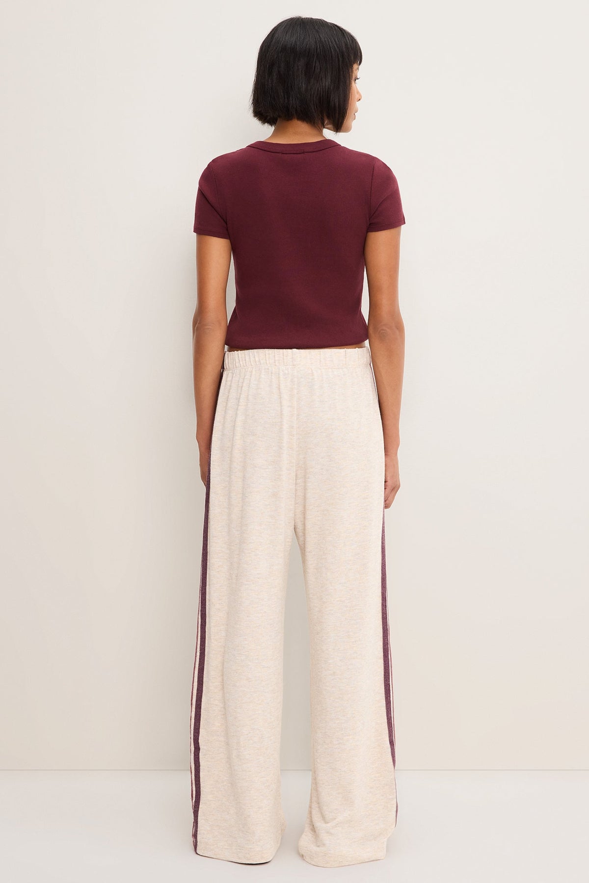 A model shown from the back wearing a short-sleeve maroon t-shirt and heather oatmeal wide-leg sweatpants with dark red side stripes, set against a plain off-white studio background.