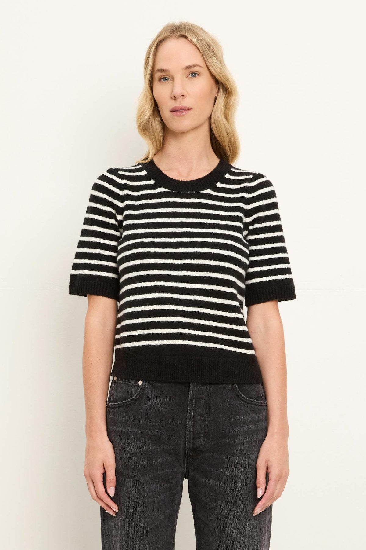 A woman modeling a short-sleeve knit sweater with horizontal black and coconut milk white stripes, paired with dark gray jeans, against a plain light background.