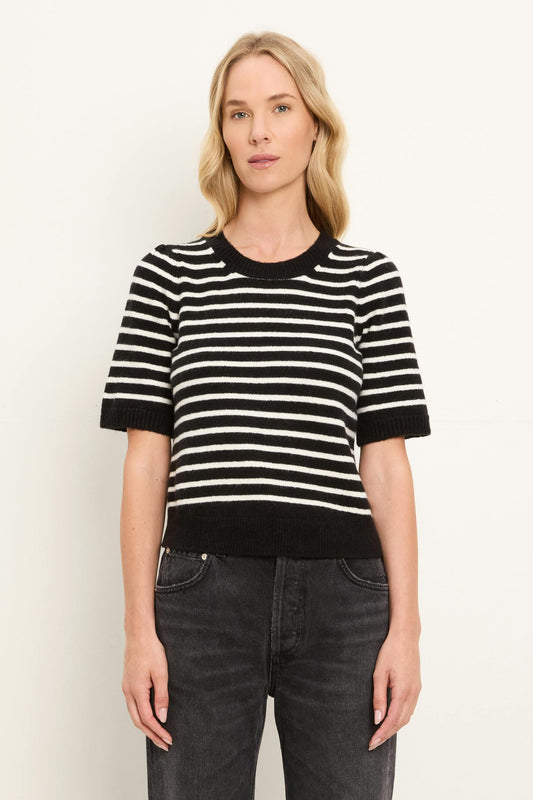 A woman modeling a short-sleeve knit sweater with horizontal black and coconut milk white stripes, paired with dark gray jeans, against a plain light background.