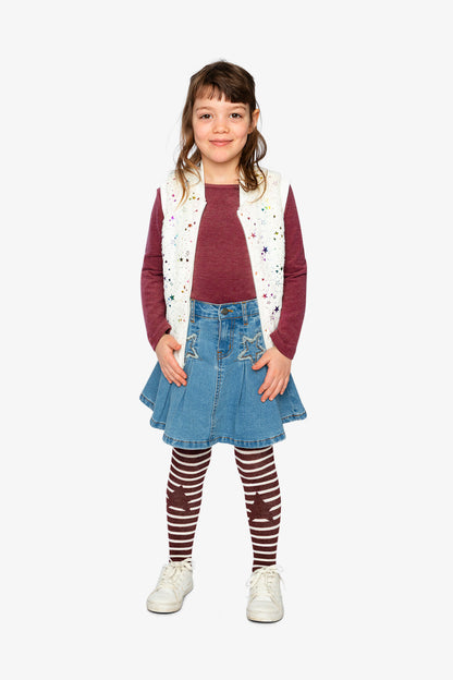 Young girl wearing a maroon sweater, with a cream stars vest over it, a denim skirt, and striped tights on a white background