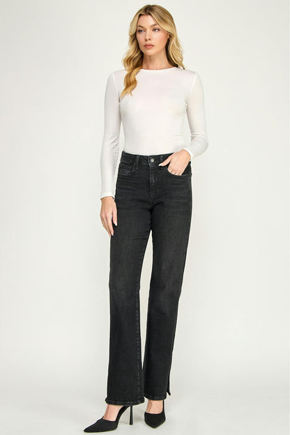 A woman modeling a white long-sleeve mock neck top and ash black high-waisted straight-leg cigarette jeans with side slits, paired with black pointed-toe heels, against a light gray studio background.