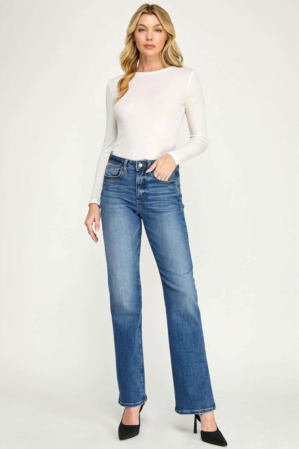 A woman modeling a pair of high-waisted, straight-leg blue jeans in baja color with a white long-sleeve top and black heels, standing against a light gray studio background.