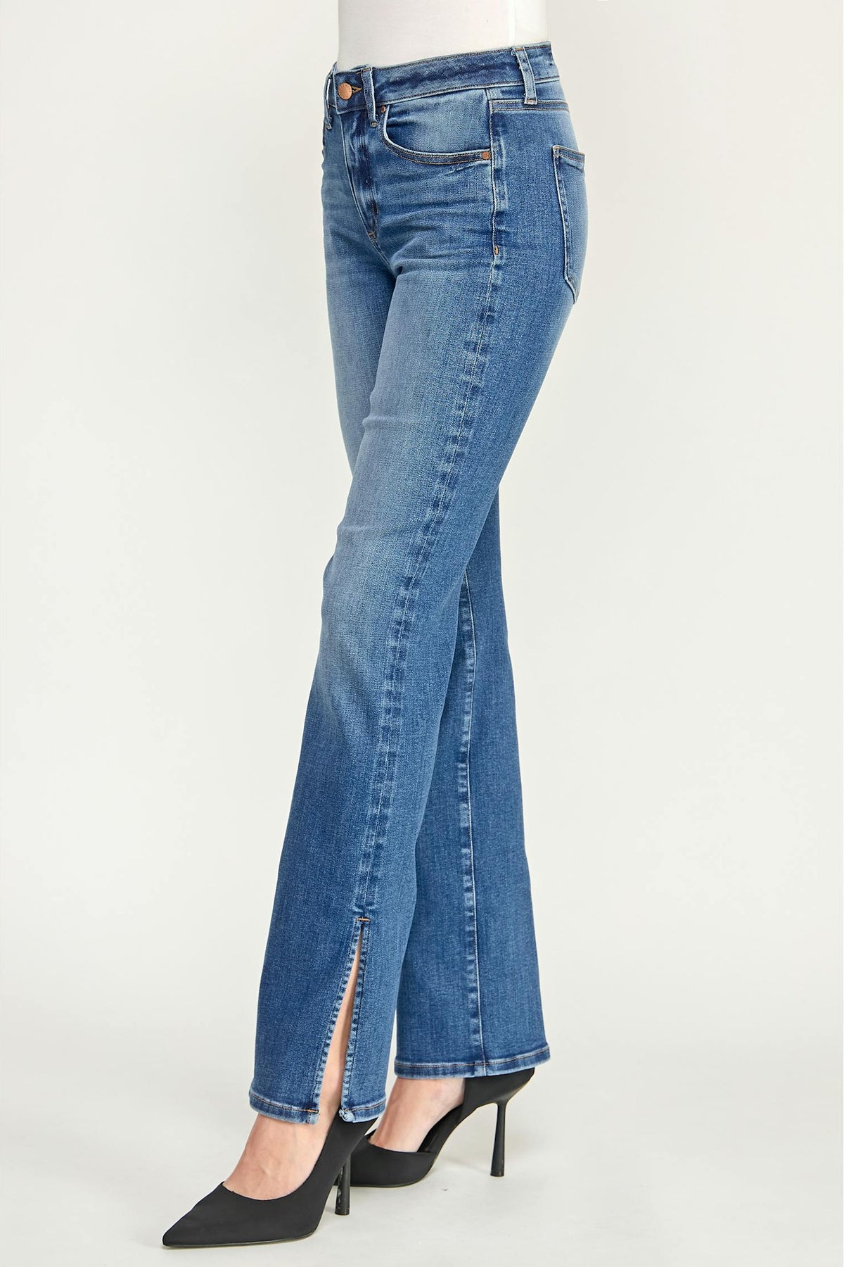 A pair of medium-wash baja blue denim  cigarette jeans with a front slit, shown on a model lower body wearing black pointed-toe heels against a plain light gray studio background.