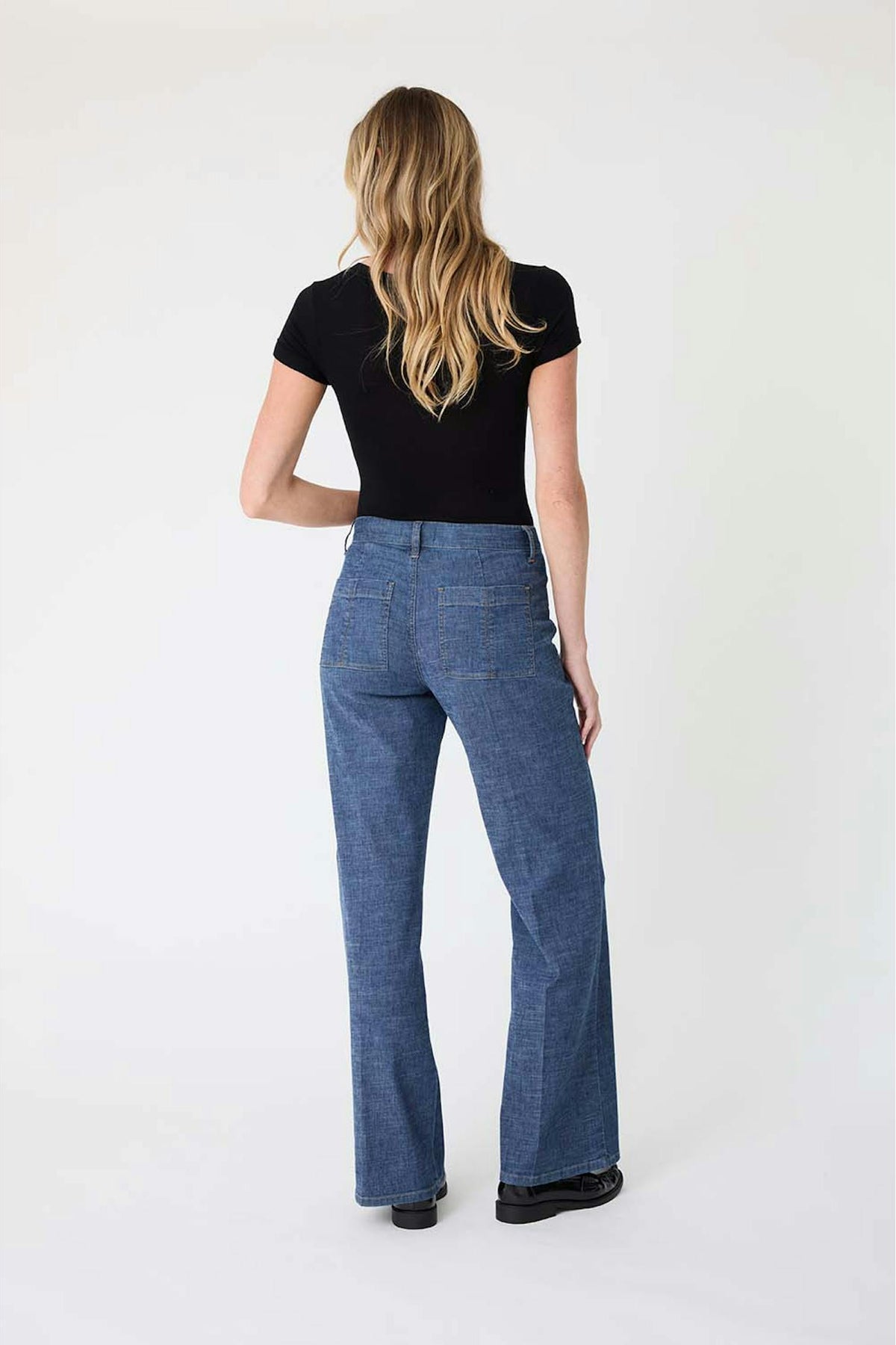 A woman viewed from the back wearing  the Sarah pant in Stargazer color, a black short-sleeve top, and black shoes against a plain light gray studio background.