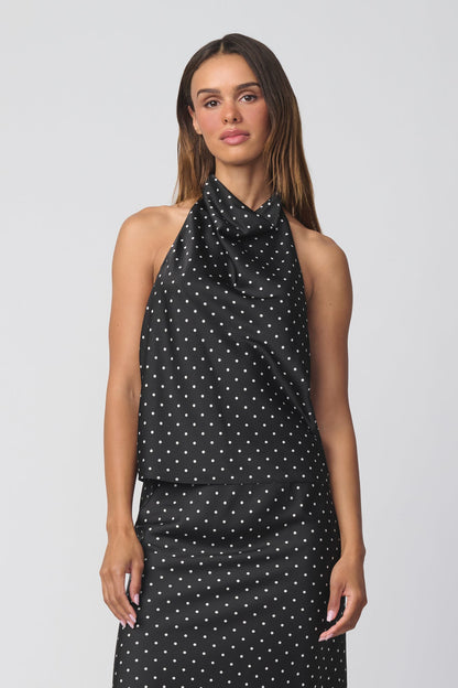 A woman modeling a sleeveless halter-neck top with a black base and small white polka dots, paired with a matching skirt and photographed against a plain light gray studio background.