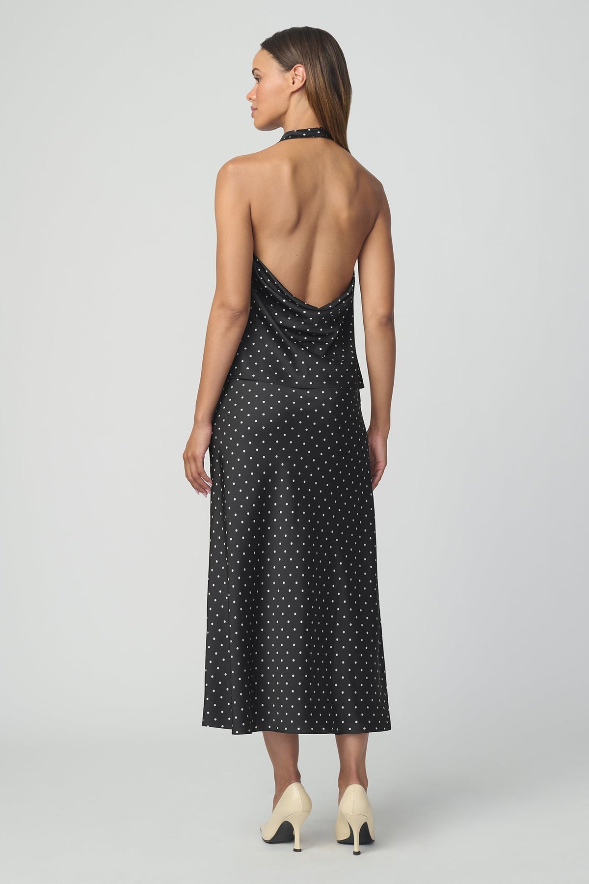 A woman shown from the back modeling a sleeveless black halter top and matching midi skirt with a white polka dot pattern  against a light gray studio background.