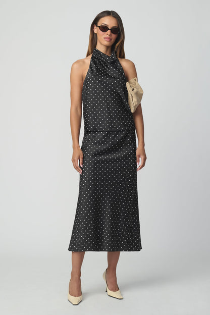 A woman modeling a sleeveless halter-neck top and matching midi skirt with a black base and small white polka dots, styled with a beige clutch and beige heels against a light gray studio background.