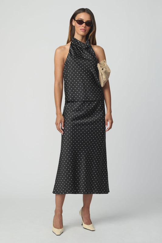 A woman modeling a sleeveless halter-neck top and matching midi skirt with a black base and small white polka dots, styled with a beige clutch and beige heels against a light gray studio background.