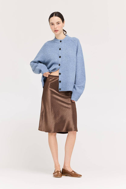 A model stands against a light beige studio background wearing a light blue cropped button-front cardigan, a chocolate brown satin midi skirt with polka dots, and brown loafers.