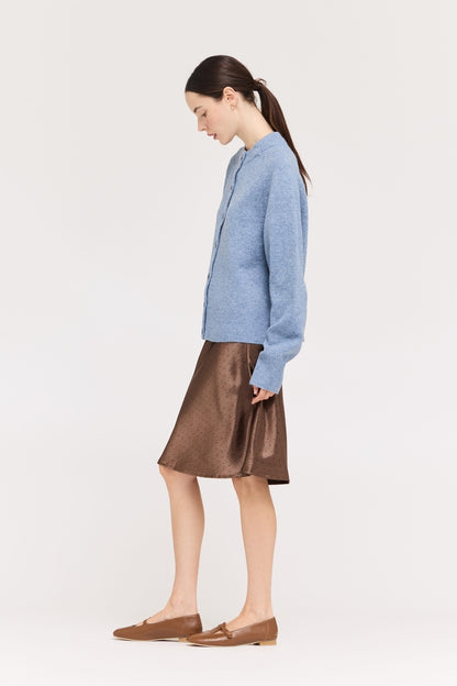 A woman in a side profile wearing a light blue knit cardigan, a chocolate brown satin A-line skirt with black polka dots, and brown leather loafers, set against a plain light gray studio background.
