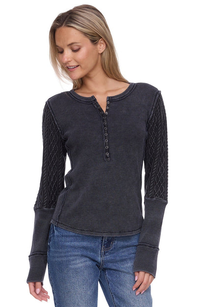A woman wearing a dark charcoal gray long-sleeve henley top with textured cable-knit sleeves, paired with blue jeans, against a plain white background.