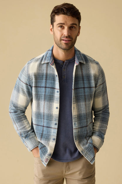 A man wearing a light blue and white plaid Asheville Vintage Wash button-down shirt over a navy blue henley t-shirt, paired with khaki pants, standing against a solid beige background.