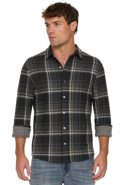A man wearing a charcoal gray and black plaid button-down shirt with rolled-up sleeves, paired with blue jeans, against a plain white background.